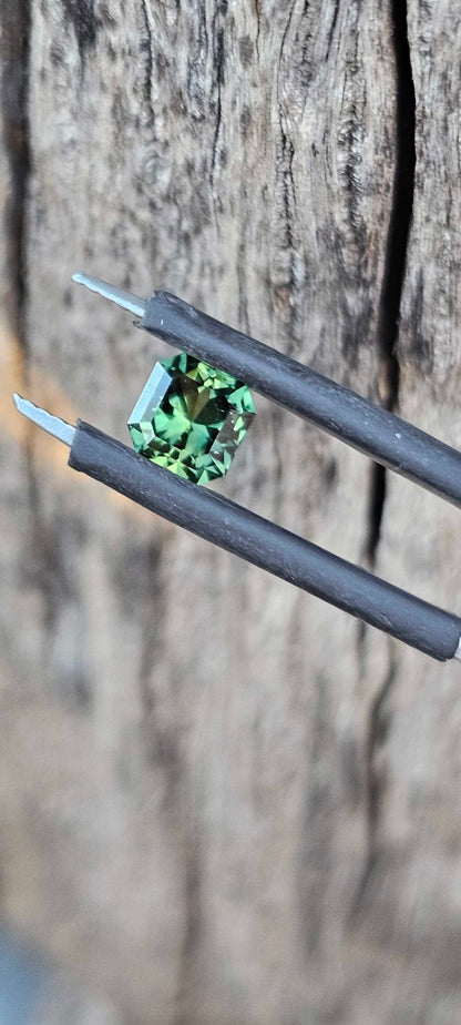 Square brilliant cut green sapphire held in jeweller's tweezers on a weathered wood background, highlighting clarity and symmetry