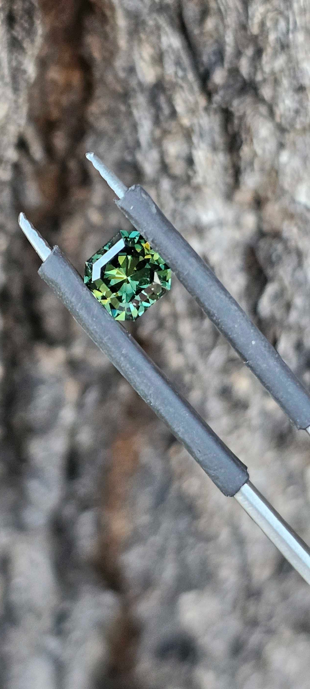 Vivid green square brilliant cut sapphire with golden undertones held in tweezers on a natural textured background