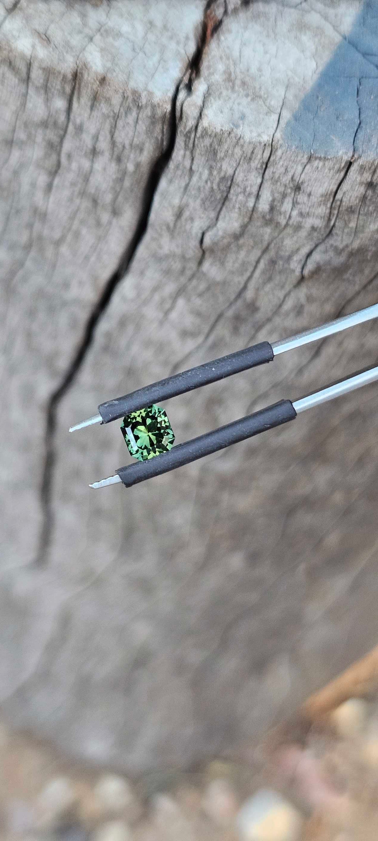 Square brilliant cut Australian sapphire photographed in tweezers over a wooden surface, showing crisp facets and bright reflections