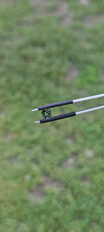Green square brilliant cut Australian sapphire held in jewller's tweezers against a soft green outdoor area